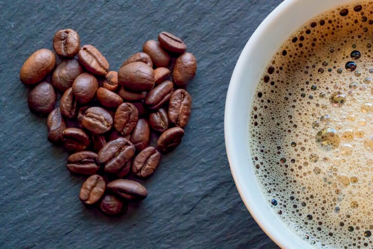 Close-up of heart-shaped coffee beans next to a cup of frothy coffee on a slate surface.